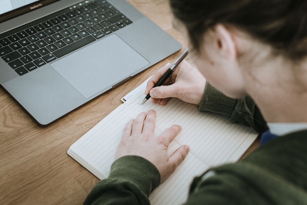 A woman writes in a notebook in front of a laptop