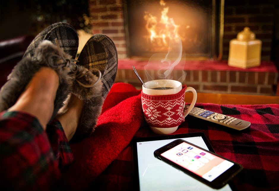 A pair of slippered feet reclines in front of a fire with a cup of steaming coffe and phone and remote next to it